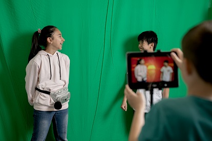 Zwei lachende Kinder stehen vor dem Greenscreen, während sie mit einem iPad gefilmt werden.